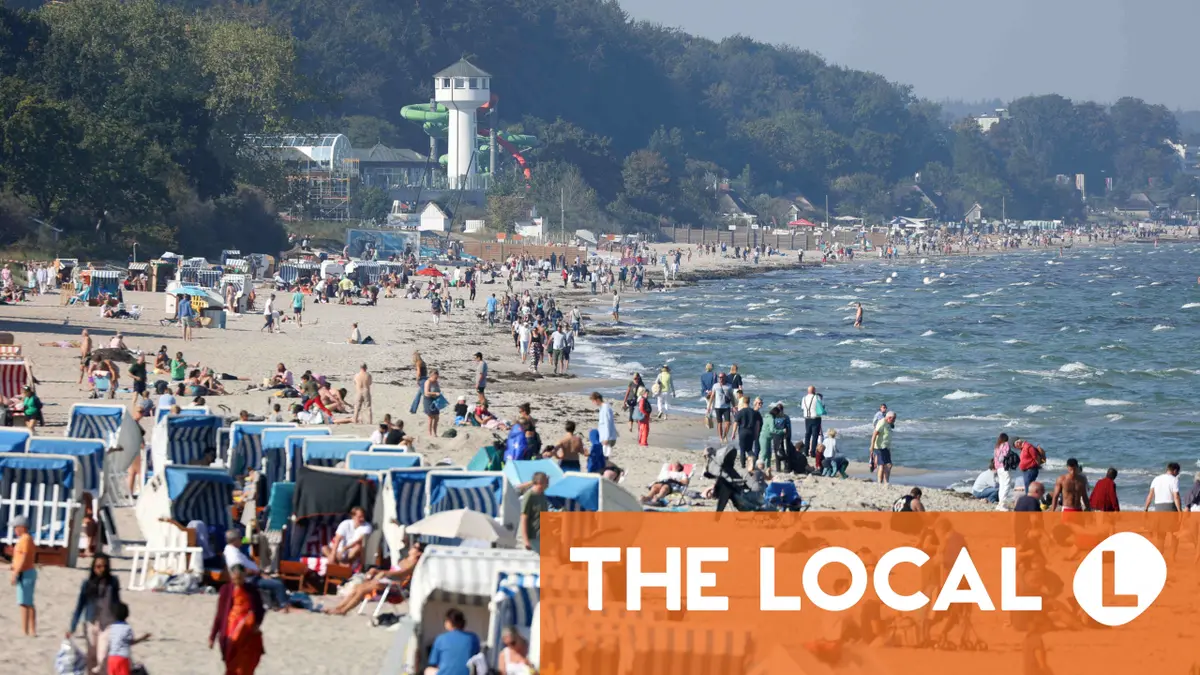 A busy beach with crowds of people, blue and white beach chairs, and a water park in the background.